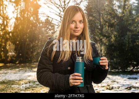 Carina ragazza bionda con capelli lunghi e piercing setto è bere tè da una tazza thermos in un parco con una certa neve e forte retroilluminazione soleggiata. Guardando a. Foto Stock