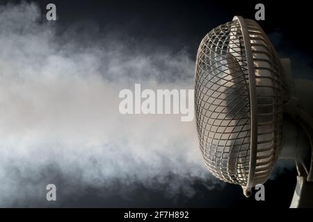 Vista dettagliata di un ventilatore da tavolo elettrico bianco su sfondo nero e pavimento riflettente con nebbia visibile o nebbia che soffia. Concetto di freschezza Foto Stock