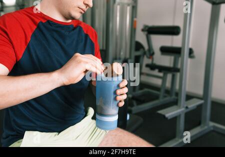 un uomo versa la proteina in uno shaker dopo un allenamento mentre si siede su una panchina in palestra. nutrizione sportiva Foto Stock