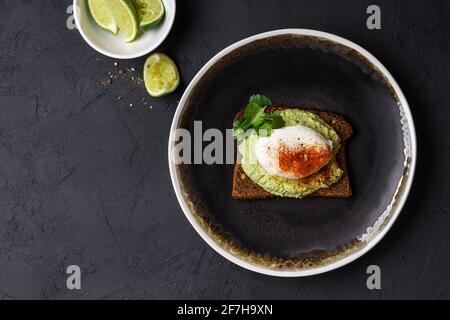 Pane di segale avocado hummus toast con uovo in camicia su piatto nero. Colazione salutare su un tavolo grigio scuro visto dall'alto, pasto o spuntino preferito Foto Stock