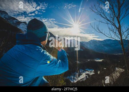 Forte flare lente con un escursionista che scatta una fotografia di una valle su montagna alpina. Il sole sta facendo un forte baluare sopra la valle alpina. Foto Stock