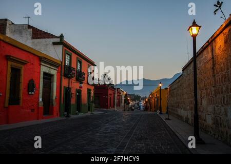 Mattina presto sulla via etipica nella città di Oaxaca, Messico. I pali della lampada sono visti sulla strada con case colorate sulla sinistra. Foto Stock