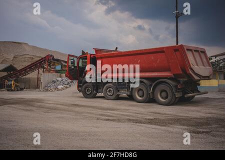 Dumptruck rosso a quattro assali parcheggiato in una cava in una giornata nuvolosa con la porta del conducente aperta. Apripista piccolo sullo sfondo. Foto Stock