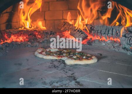 Pizza fatta in casa in forno caldo, panificio italiano vecchio stile per pizza a casa. Tronchi e fuoco visto dietro una deliziosa pizza in primo piano Foto Stock