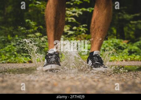 Le gambe maschili saltano in un piccolo stagno sul asfalto, creando un grande spruzzi di acqua chiara che sta uscendo da sotto scarpe sportive che l'uomo sta indossando Foto Stock