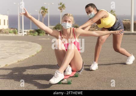 Due amici femminili con maschere protettive che giocano con skateboard. Una ragazza che spinge l'altra da dietro. Concetto di amicizia e coronavirus. Foto Stock