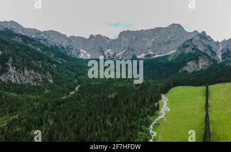 Panorama aereo di campo verde e cottage sotto le montagne alpine in una giornata di sole nelle Alpi Giulie, Slovenia, guardando verso la vetta alpina dell'uomo Foto Stock