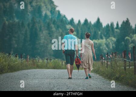 Una coppia appena sposata si allontana dalla macchina fotografica su una strada sterrata con una recinzione e una lussureggiante foresta sullo sfondo. Foto Stock