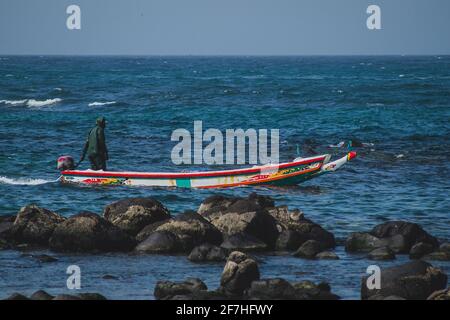 Tipica barca da pesca in Almadies, Dakar, Senegal, chiamato piroga o piragua o piraga con un pescatore che guida la barca in una baia su una chiara da soleggiata Foto Stock