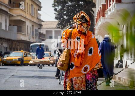 Vista posteriore di una tipica donna africana vestita di colorati abiti arancioni per le strade di Sant Louis, Senegal durante mezzogiorno. Foto Stock