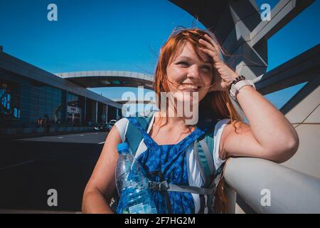 Una giovane donna che viaggia con capelli rossi, che porta due zaini e una bottiglia d'acqua, si appoggia sulla recinzione e gode del sole fuori dall'aeroporto. Foto Stock