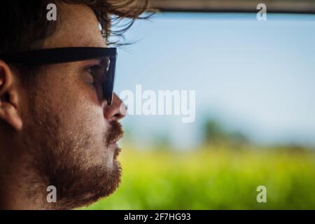 Immagine laterale o di profilo di un giovane uomo che guida un'auto con la finestra aperta che espone i campi verdi e il cielo blu. Immagine effetto vintage con ret Foto Stock