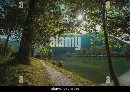 Tranquillo lago del fiume Soca al massimo na Soi in mattina presto. Vista tra gli alberi verso un sentiero in ghiaia accanto al lago. Foto Stock