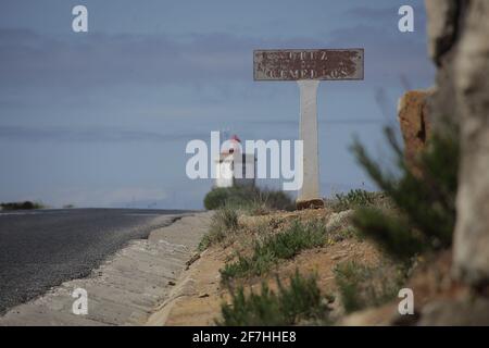 Un cartello in cemento usurato accanto a una strada, con un faro bianco sullo sfondo in una giornata di sole a Peniche, Portogallo. Foto Stock