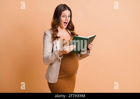 Foto di dolce donna impressionata che aspetta il bambino vestito marrone libro di lettura tummy isolato beige sfondo di colore Foto Stock