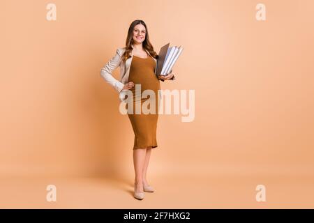 Foto di dolce adorabile donna che aspetta il bambino vestito marrone pancino che tiene le cartelle isolato sfondo di colore beige Foto Stock