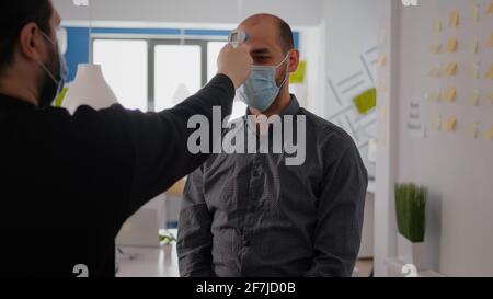 Uomo freelance che indossa una maschera protettiva contro il covid19 mentre controlla la temperatura utilizzando un termometro medico per evitare l'infezione da coronavirus. Azienda che prende precauzioni durante la pandemia globale Foto Stock