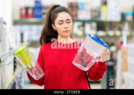 Ritratto di giovane donna sceglie tra due caraffe con un filtro dell'acqua in un negozio di elettrodomestici. Il concetto di consumismo. Foto Stock