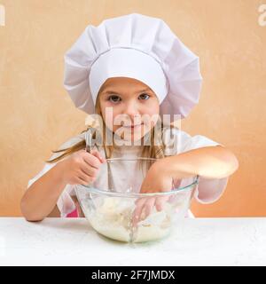 Una bambina vestita come cuoco impasta l'impasto. Cucina bambino stile di vita concetto. Il capretto ama, ha divertimento, studia e gioca nella cucina Foto Stock