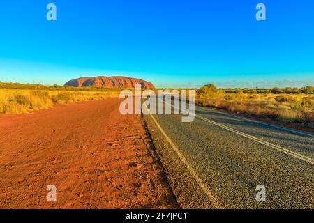 Strada che conduce a Uluru Ayers Rock a colori vibranti del tramonto. L'enorme monolito di arenaria icona dell'entroterra australiano nel Parco Nazionale di Uluru-Kata Tjuta Foto Stock