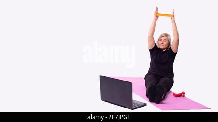 Banner, long format.handsome donna anziana che guarda l'allenamento online su computer portatile e utilizzando una fascia elastica in corda durante l'allenamento mattutino, sfondo grigio Foto Stock