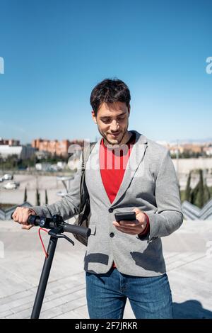 Foto in stock di un uomo d'affari in giacca con zaino stare in piedi su uno scooter elettrico e utilizzare lo smartphone Foto Stock