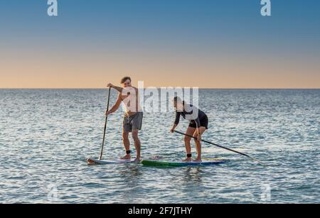 Coppia caucasica matura su SUP paddleboarding divertendosi su un mare tranquillo al tramonto in una bellissima spiaggia remota. Persone anziane moderne attive, all'aperto A. Foto Stock