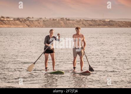 Coppia caucasica matura su SUP paddleboarding divertendosi su un mare tranquillo al tramonto in una bellissima spiaggia remota. Persone anziane moderne attive, all'aperto A. Foto Stock