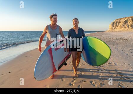 Coppia matura con tavole da surf sulla bellissima spiaggia godendo di paradiso e di uno stile di vita attivo. Bella forma uomo e donna surf e divertirsi. In trave Foto Stock