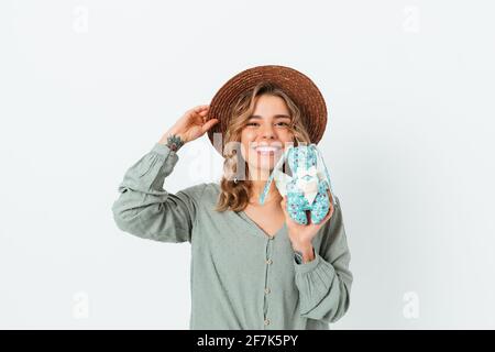 Allegra ragazza bionda contenta che tiene il suo cappello di paglia e mostra coniglio giocattolo mentre si trova su sfondo bianco studio. Foto Stock