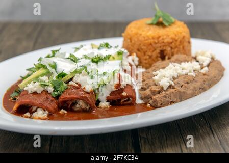 Enchiladas di pollo affumicato in salsa rossa servita con fagioli e riso fritti per un gustoso pasto messicano. Foto Stock