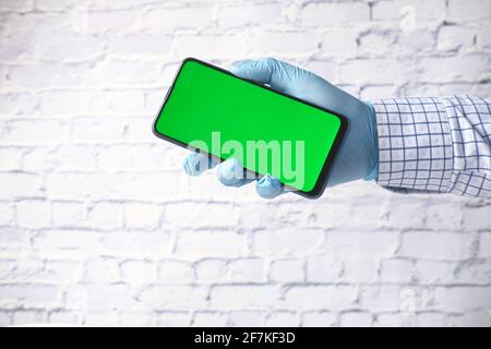mano del medico in guanti protettivi che tengono lo smartphone con verde schermo Foto Stock