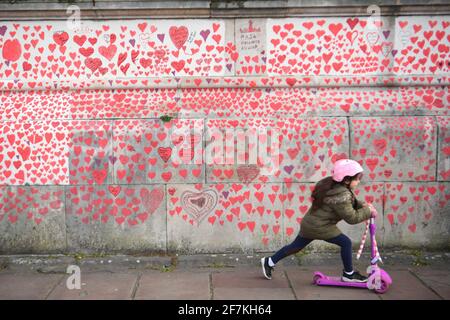 Una giovane ragazza cavalcava uno scooter davanti al National Covid Memorial Wall sull'argine di Londra. Più di mille persone, tra cui il personale di NHS, i volontari e i membri del pubblico in lutto, hanno dipinto 150,000 cuori sul muro, che ora si estende per circa mezzo chilometro, per rappresentare i cari persi a causa del virus nel Regno Unito. Data immagine: Giovedì 8 aprile 2021. Foto Stock