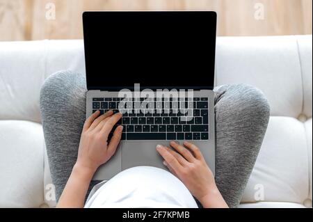Concetto di tecnologia, gadget wireless. Vista dall'alto di una donna incinta in una t-shirt bianca seduta su un divano, tenendo un computer portatile con schermo nero vuoto in mano, posto per la vostra presentazione Foto Stock