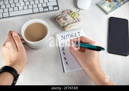 Vista dall'alto delle mani femminili che scrivono in blocco note, nota con le parole BUDGET, tazza di caffè e denaro sul tavolo. Concetto di mentalità del costruttore di ricchezza, distribuzione Foto Stock