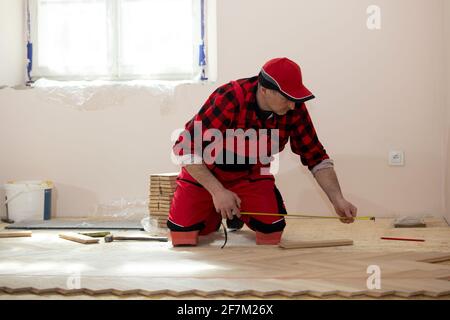 Uomo costruttore che misura per l'installazione del pavimento in legno nella ristrutturazione di una casa. Falegname che installa pavimenti in laminato in camera Foto Stock