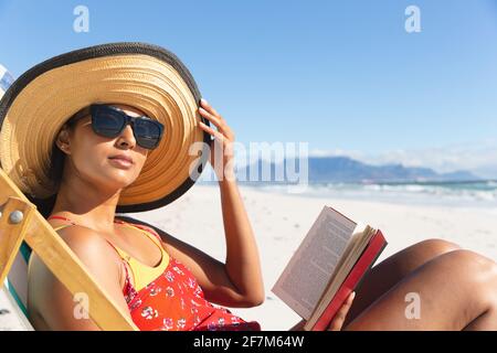 Razza mista donna in vacanza sulla spiaggia seduta in sdraio lettura prenota Foto Stock