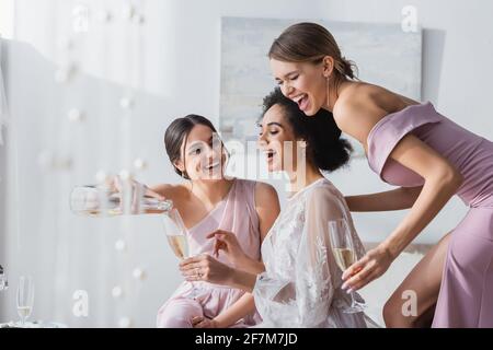 giovane donna che versa champagne vicino afroamericana sposa ed eccitata sirena Foto Stock