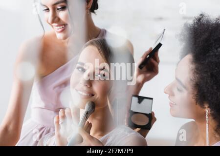 sposa felice guardando la macchina fotografica mentre le donne interracial che applicano il trucco, primo piano offuscato Foto Stock