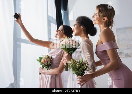 sposa afroamericana con bridesmaid che tiene bouquet di nozze mentre prende selfie sullo smartphone Foto Stock