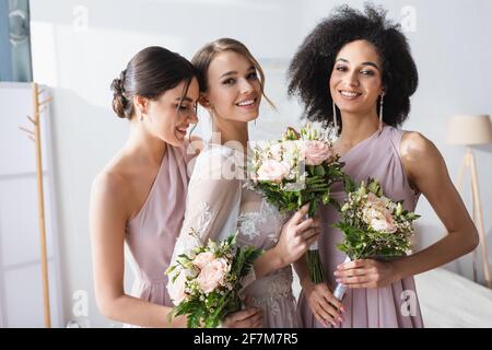 bella sposa insieme con bridesmaids interracial che tiene i bouquet di nozze dentro camera da letto Foto Stock