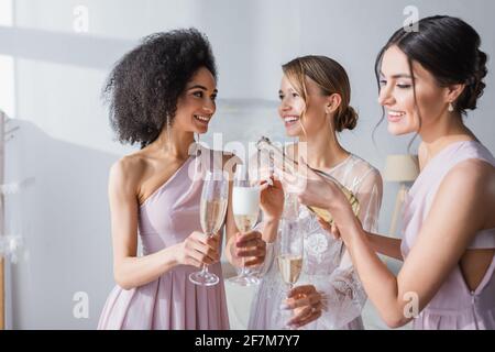 donna sorridente che versa champagne vicino a sposa felice e afroamericana sirena Foto Stock