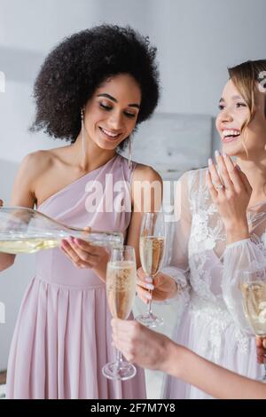 elegante donna afroamericana versando champagne vicino sposa felice Foto Stock