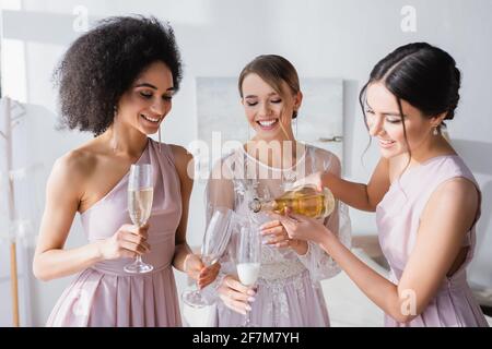 bella donna versando champagne vicino sposa e afroamericana bridesmaid Foto Stock