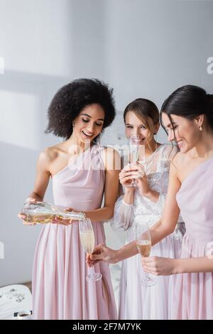 donna afroamericana che versa champagne vicino a bride allegra e bridesmaid Foto Stock