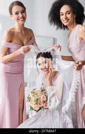 bridesmaids interrazziali felici sorridendo alla macchina fotografica mentre tiene il velo sopra sposa giovane Foto Stock