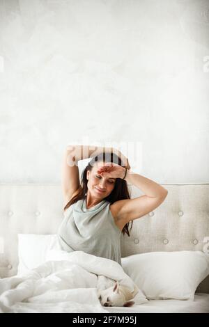 Concetto di mattina pigra. Bella donna felice si sveglia a letto e striscia le mani. Bella donna che riposa a casa in camera da letto. Il cane piccolo dorme vicino Foto Stock