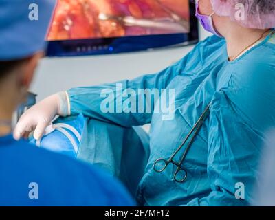 Messa a fuoco selettiva su un morsetto chirurgico su un abito blu di un medico in una sala operatoria chirurgica di fronte a un monitor. Foto Stock