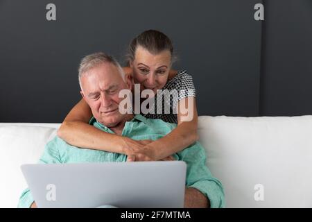 Felice coppia caucasica senior in soggiorno guardando il computer portatile abbracciare e sorridere Foto Stock