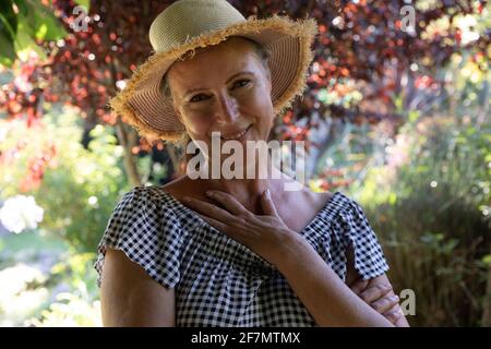 Ritratto di felice donna caucasica anziana in piedi in giardino soleggiato con cappello, sorridente alla macchina fotografica Foto Stock